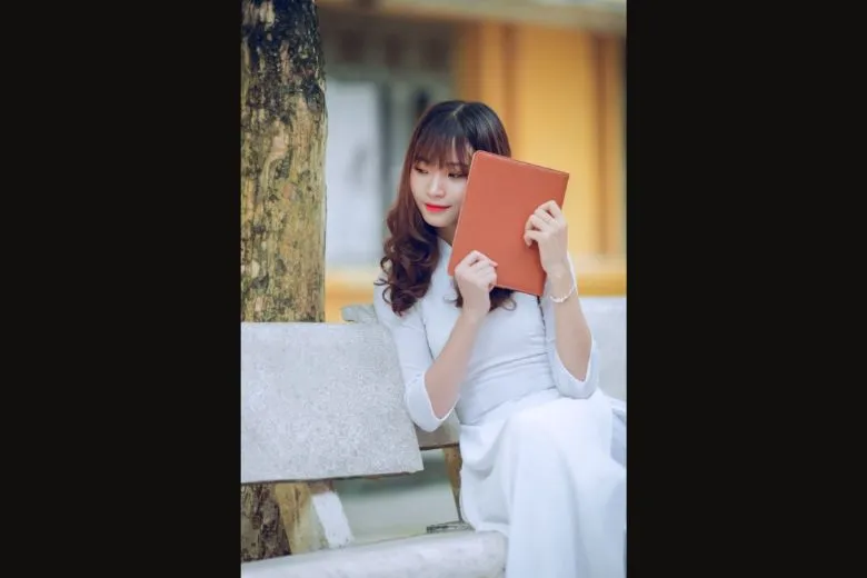 Nữ sinh áo dài trắng ngồi ghế đá, tay cầm nón lá